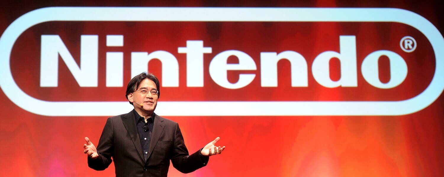 Satoru Iwata Passes Away Satoru Iwata standing in front of the Nintendo logo.