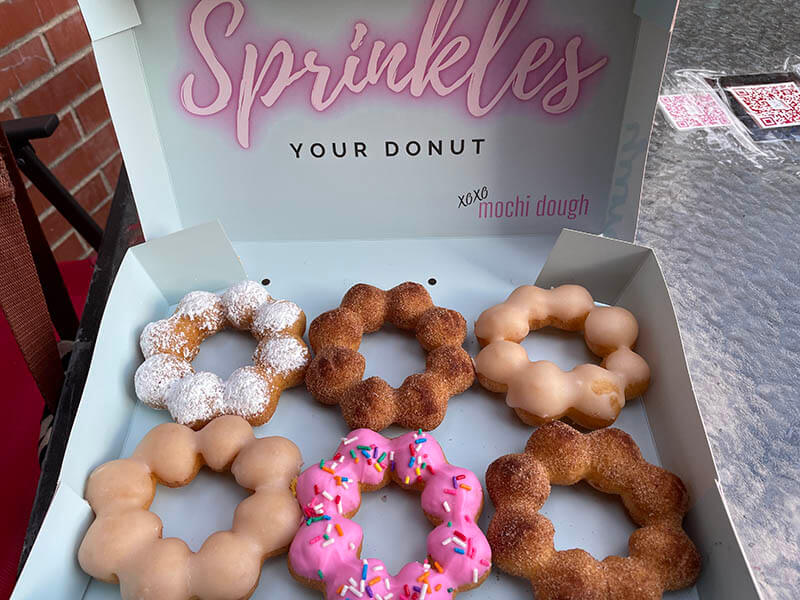 6 various mochi donuts in a box that says, "Sprinkles your donut. XOXO Mochi Dough."
