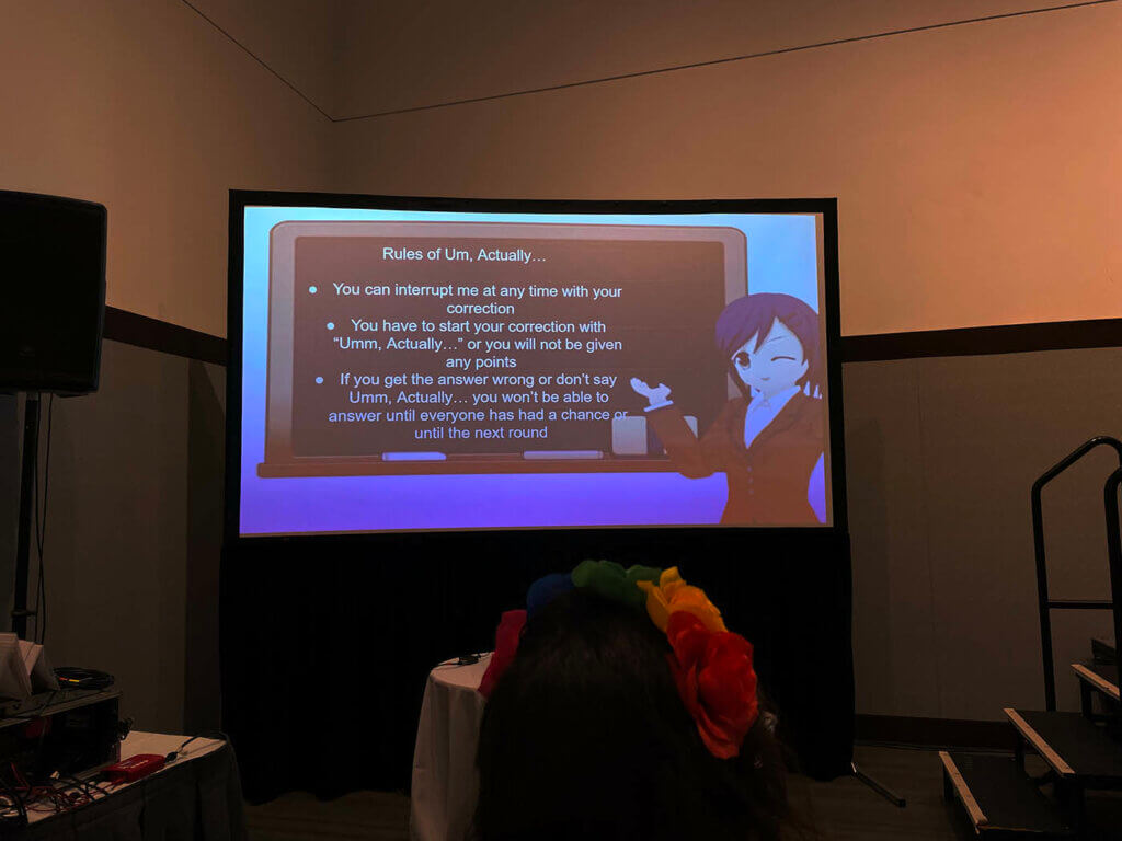 The Um, Actually Rules projected on the screen with an anime character presenting them. Rules of Um, Actually... "You can interrupt me at any time with your correction. You have to start your correction with 'Um, actually...' or you will not be given points. If you get the answer wrong or don't say 'Um, actually'... you won't be able to answer until everyone has had a chance to answer or until the next round."