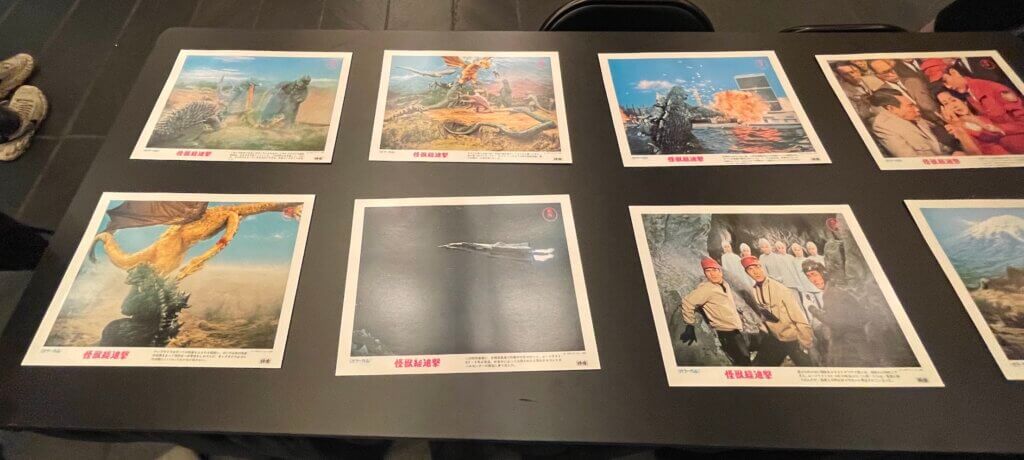 There is a table with 7 movie posters from Destroy All Monsters on display. Some of the scenes depicted include the spaceship the human characters used, the final final scene between the monsters, and the actors.