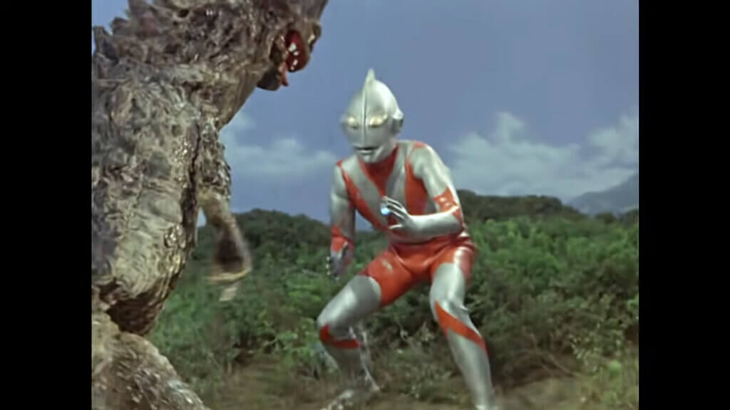 Ultraman faces off against a lizard-like kaiju with a gaping toothy maw and scaly, warty skin. they are standing in a dry field bordered by green brush.