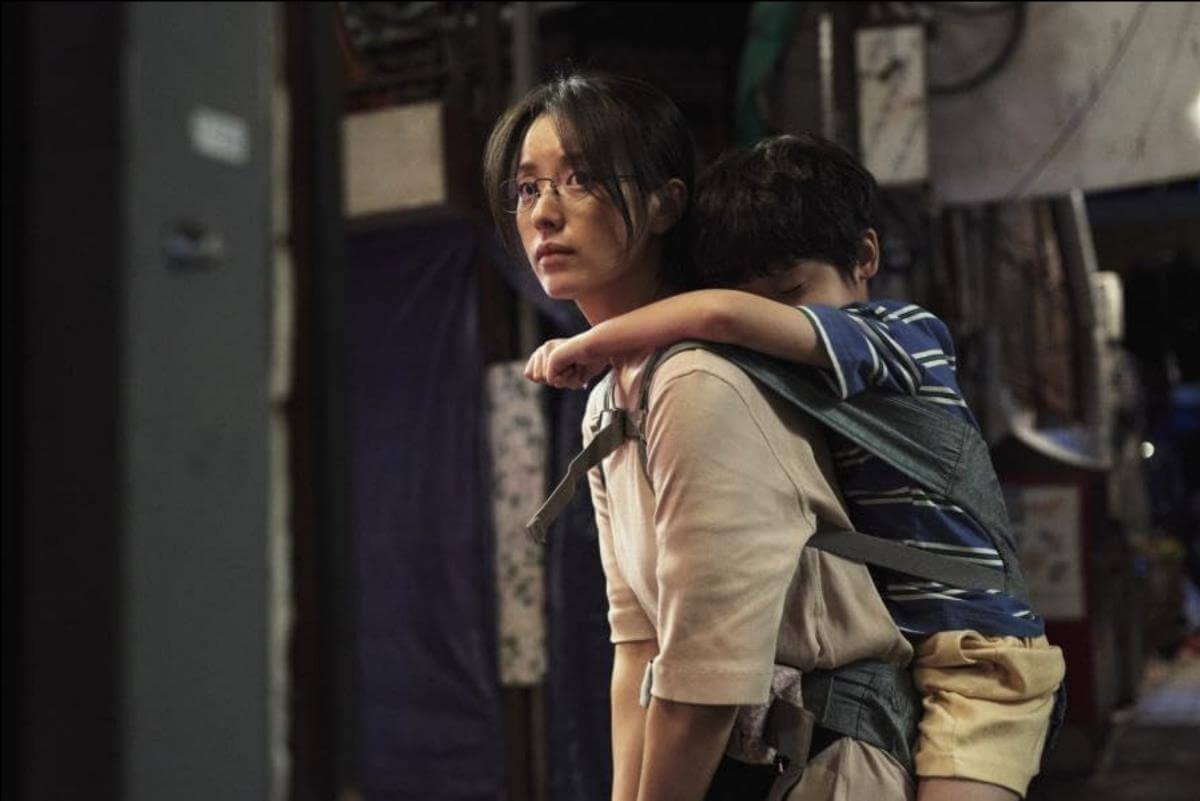 Footage from the TV series Moving. A young woman wearing a white shirt and glasses carries a child via a sling on her back. The child has his arms around her neck. He is wearing a blue and white striped shirt and khaki shorts. Behind them is the back alley of a Korean city. 