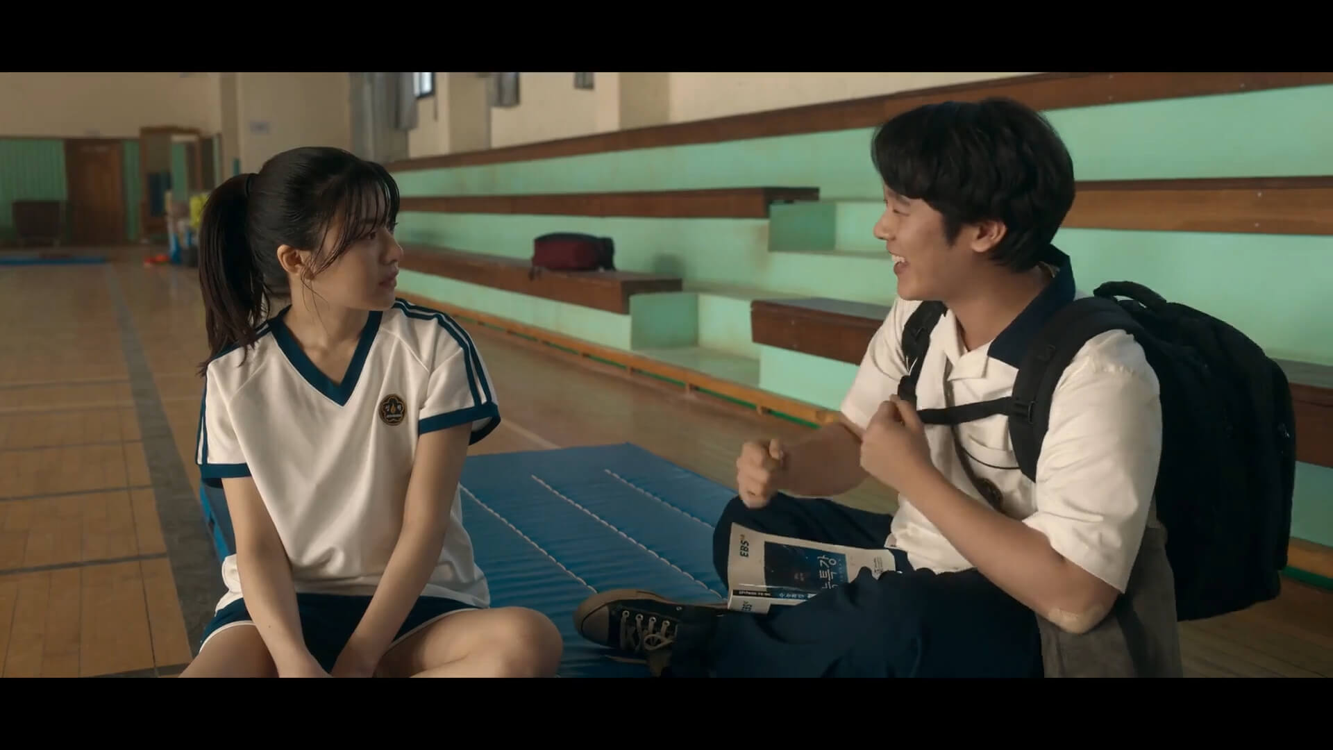 A sequence from the TV series "Moving." In a high school gymnasium, a teenage boy wearing a heavy black backpack shadowboxes while sitting and smiling. A teenage girl looks at him with a befuddled expression on her face. Both are wearing white gym uniforms.