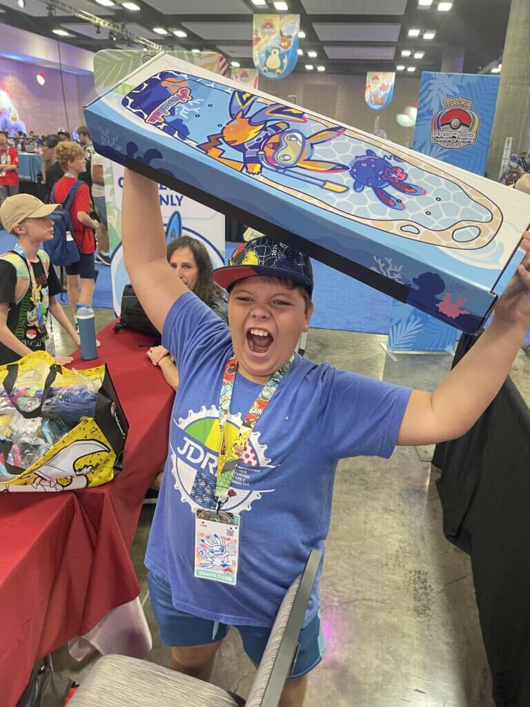 A young boy with a competitor badge around his neck excitedly holds up the exclusive Bear Walker skateboard above his head. The skateboard has Pikachu in a scuba diving outfit swimming underwater with an Azumarill swimming above it.