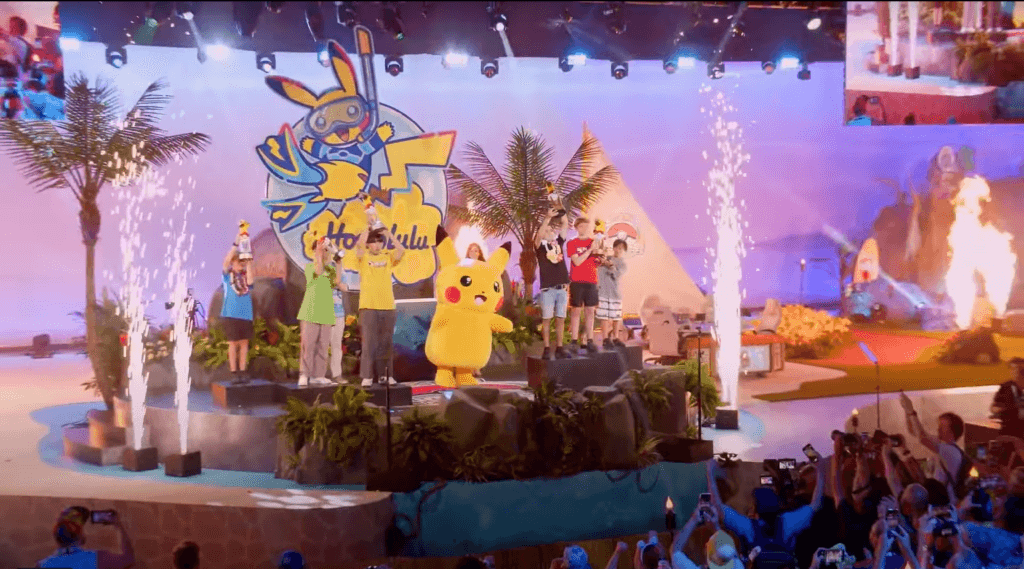 The TCG juniors, seniors, and masters winners and finalists are standing on podiums, lifting their trophies into the air while fireworks go off around the stage and a person in a pikachu costume dances in the center.