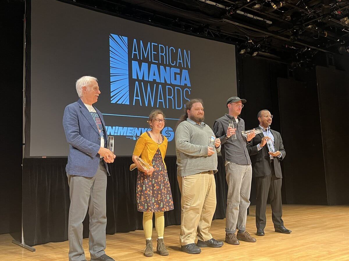 Fred, Lys, Thomas McAlister, Adam Grano and Ajani Oloye stand on stage. From left to right: older man with mustache, woman with yellow jacket and flower dress, man with long curly hair in ponytail, tall skinny man wearing baseball cap, man with buzzed hair wearing black suit and pants.