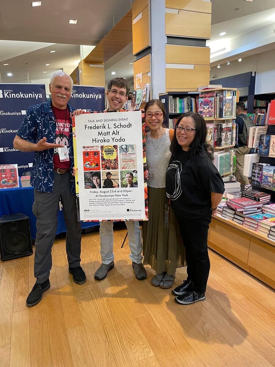 Fred Schodt, Matt Alt, Hiroko Yoda and Deb Aoki stand together in Kinokuniya holding a sign displaying their photos and book covers. There are book shelves behind them. From left to right: older man, man wearing glasses, woman wearing necklace and glasses, woman wearing tshirt and glasses