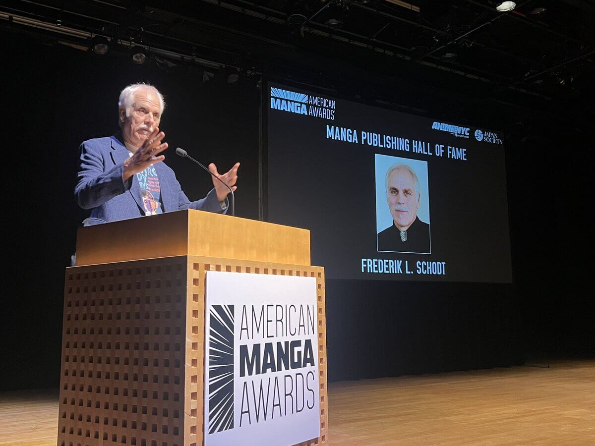 Fred Schodt, an older man wearing a suit, lectures at the American Manga Awards podium. A photograph of himself is projected on the screen behind him.