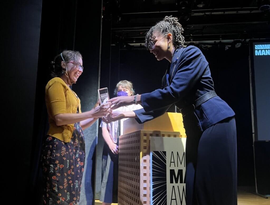 Lys Blakeslee, wearing a yellow jacket on the left, receives the award from a woman on the left wearing a blue jacket on stage at the American Manga Awards. Another woman stands at the podium behind them.