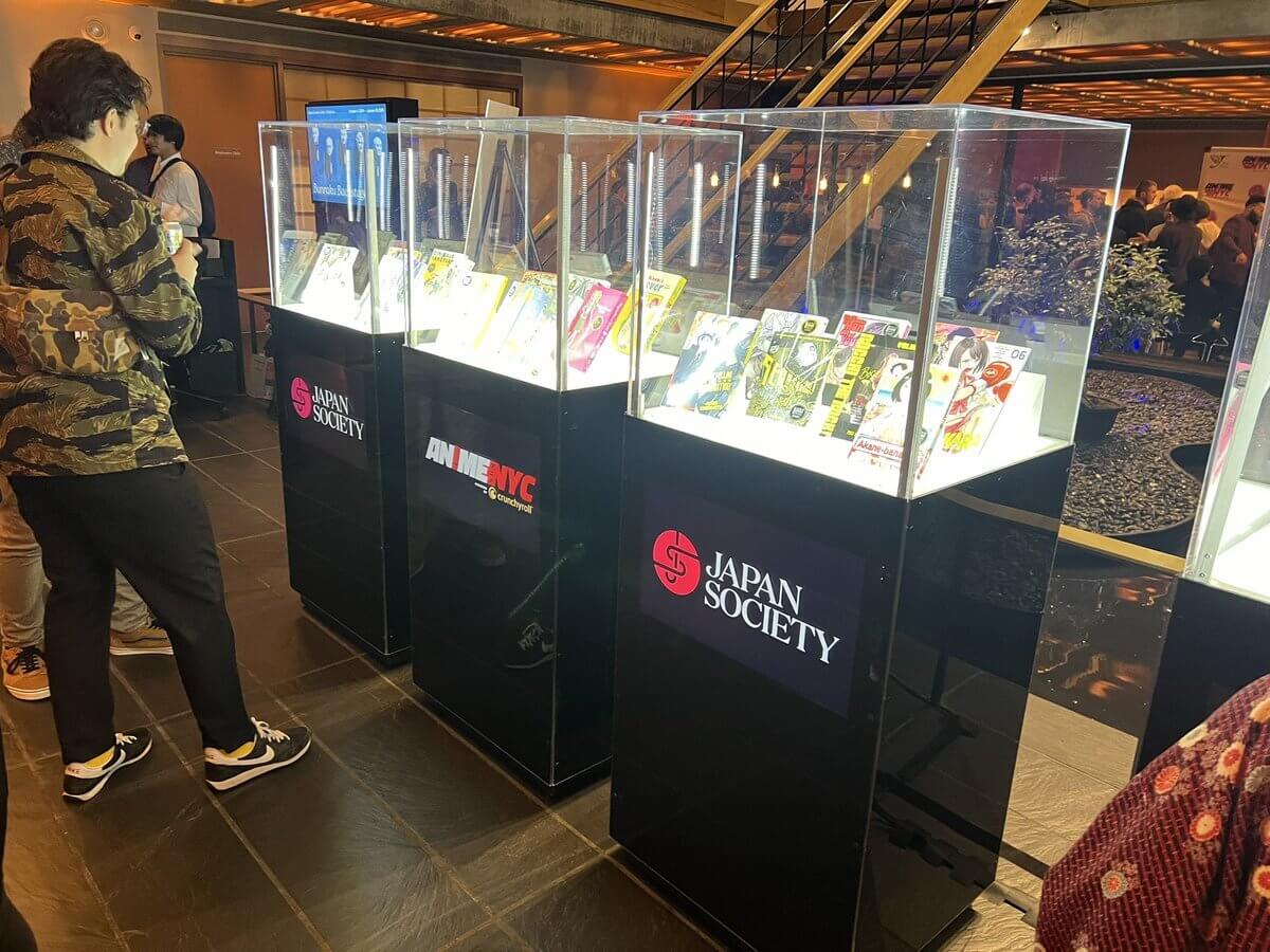 Display cases exhibiting nominated books at the American Manga Awards. Casess are marked with Japan Society logo. Somebody is taking pictures in the upper left corner of the image.