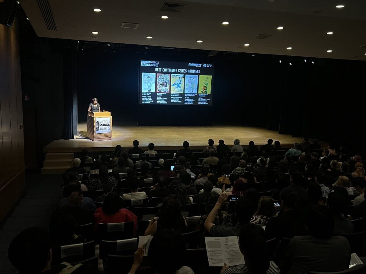 Woman at podium presents Best Continuing Series on stage at the American Manga Awards. The covers of these series are projected on the screen to her right. The picture is taken from the back of the audience.