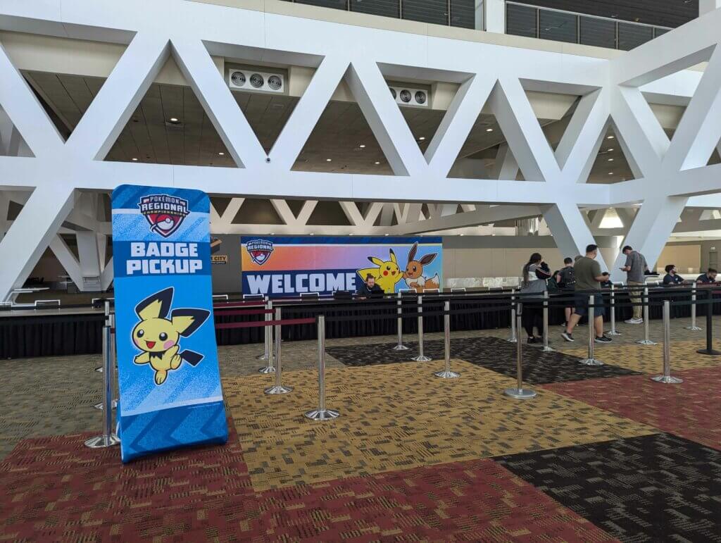 The Badge Pickup line with a sign showing Pichu smiling and showing the way to register. Behind the counter is a a large welcome sign with a Pikachu and an Eevee high-fiving.