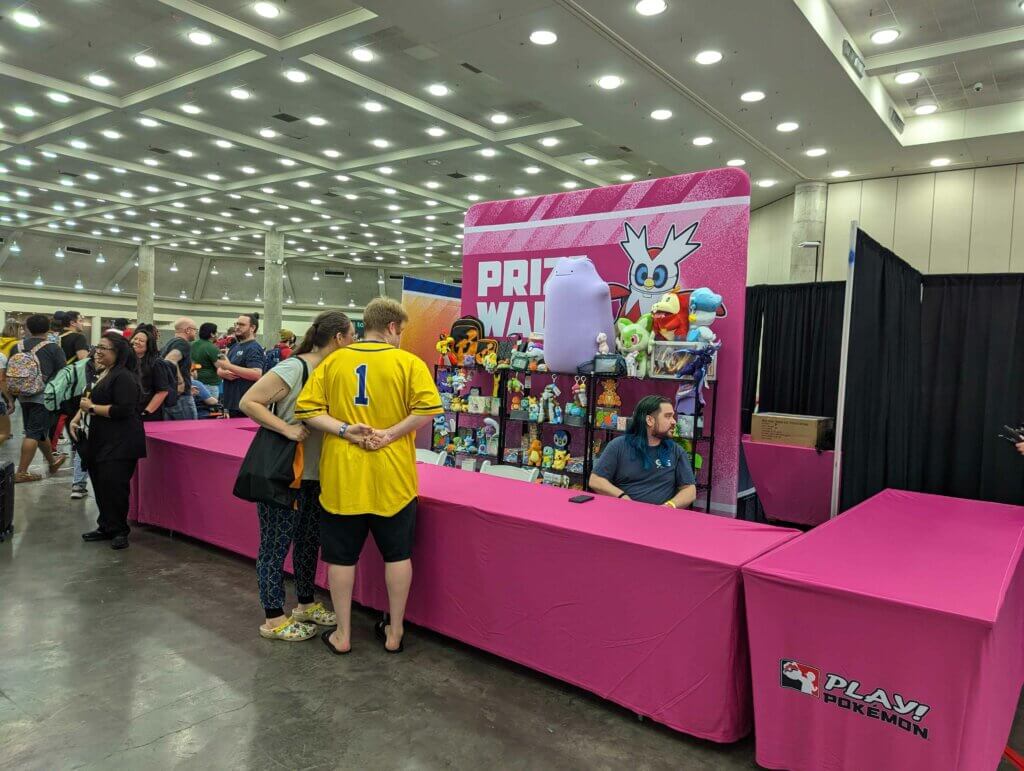 The prize wall is decorated in pink with a Delibird on the sign and a few shelves set up to showcase a small variety of plushies, bags, and card packs available for purchase using side event prize tickets.