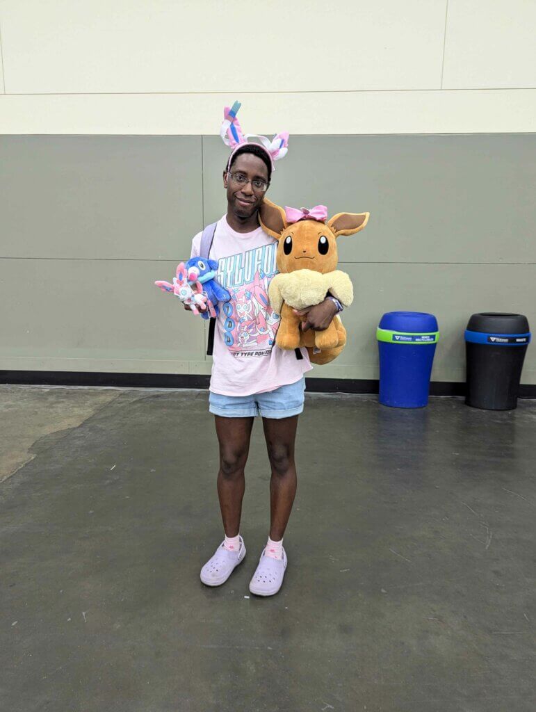 A Pokémon fan wearing a pink shirt with Sylveon on it, a headband giving them Sylveon ears, and matching pink socks and purple shoes. They’re carrying a large Eevee plush with a pink bow on its head as well as smaller Sylveon and Poplio plushies.