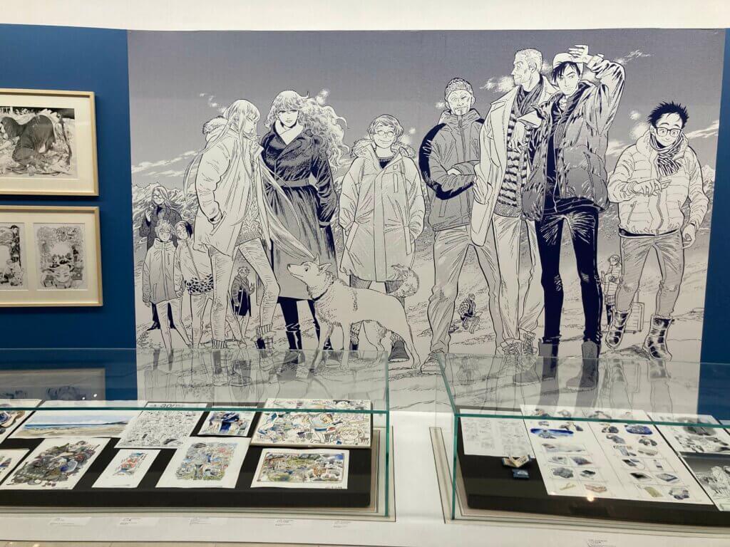 Irie’s colored art rests under a glass case. Above is a blown out panel from Go With the Clouds North-by-Northwest, featuring several women and men together with a dog standing on the ice.