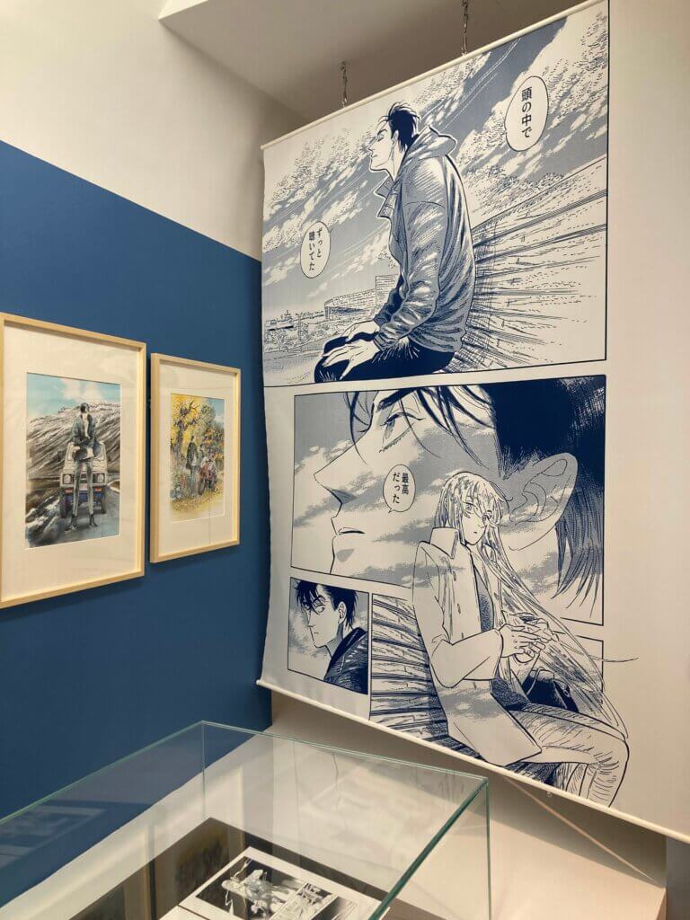 A scene from the Aki Irie section of the exhibit. To the right of a glass case featuring Irie’s illustrations, a page from Go With the Clouds North By Northwest is blown up on a hanging scroll. To the left are framed and mounted color Irie illustrations.