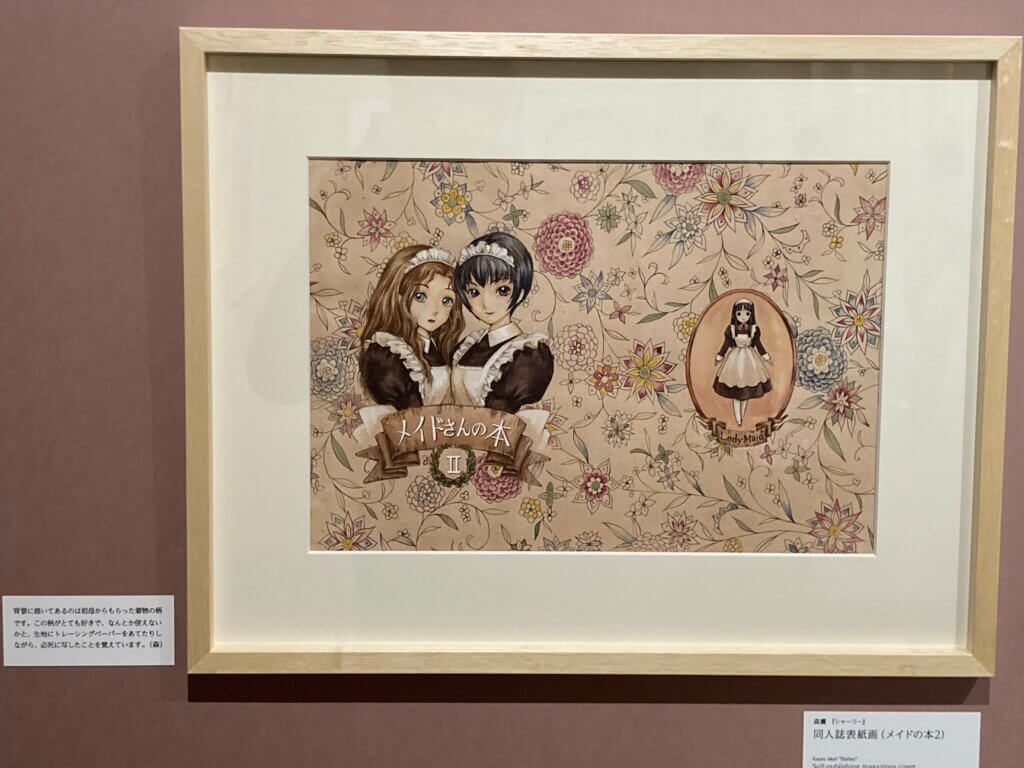 The framed cover design of a Kaoru Mori dojin comic, “Maid-san no Hon II” or “Maid Book II.” Two women pose together wearing maid costumes against a flower print background. A long-haired maid poses to the right within a labelled oval.