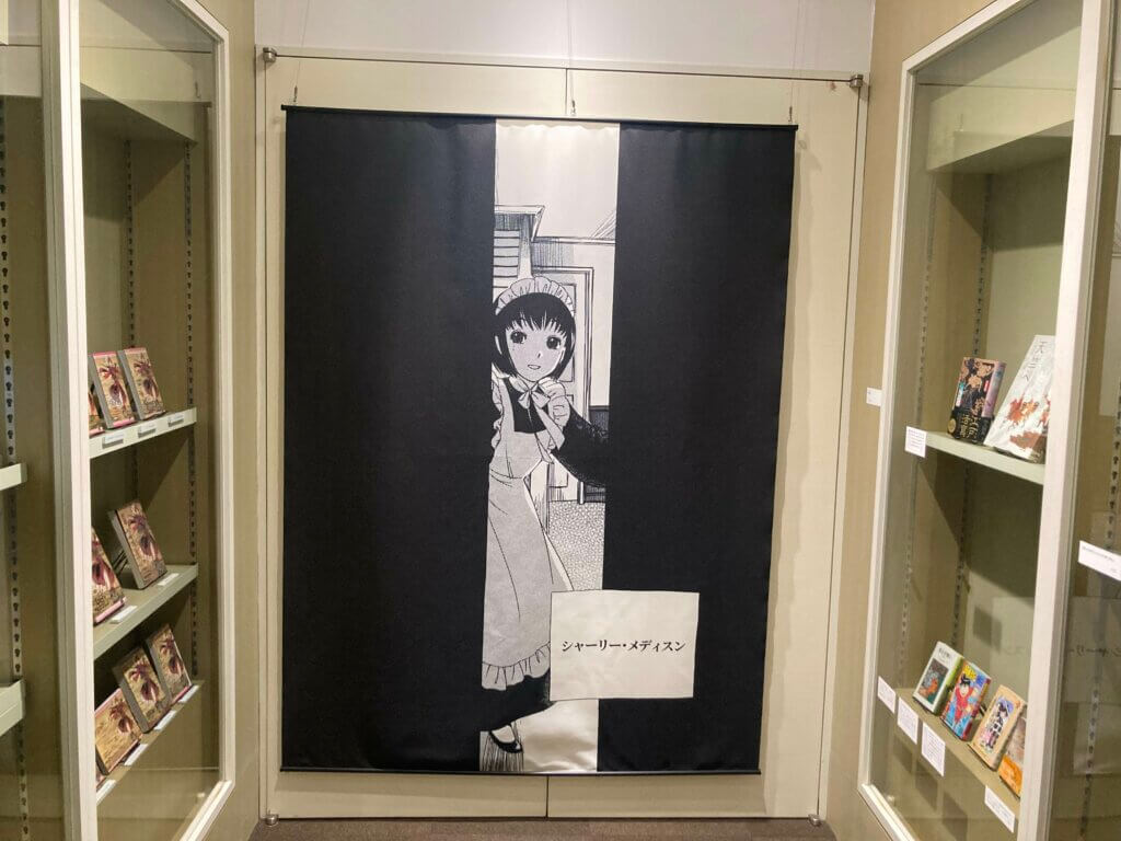 Nook of books featuring various translated editions of A Bride’s Story’s [italics] first volume, as well as recommended titles from Aki Irie. At the end is a wall hanging depicting a young woman with short black hair wearing a maid costume as seen through a door that she is opening.