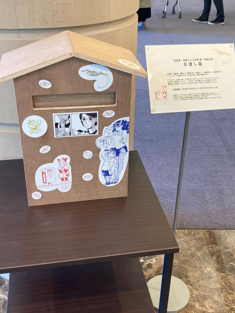 A wooden postbox sits on a table outside the exhibit. It is meant to store messages for the manga artists Kaoru Mori and Aki Irie. The box is covered with stickers depicting characters from Irie’s Ran and the Grey World and Mori’s Emma. 