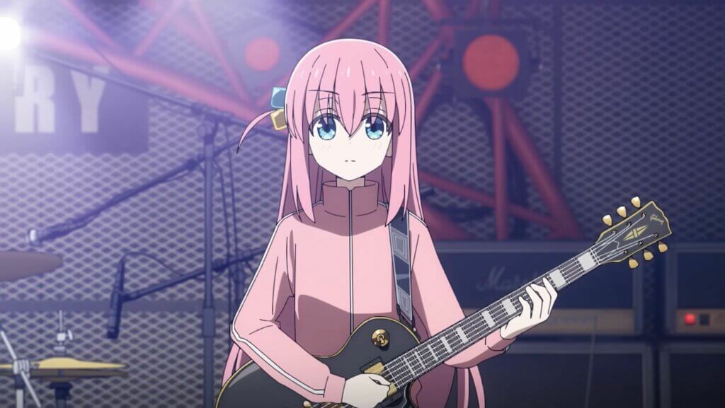 Image is Bocchi, a long pink haired girl with blue eyes wearing a pink tracksuit and holding a black electric guitar.