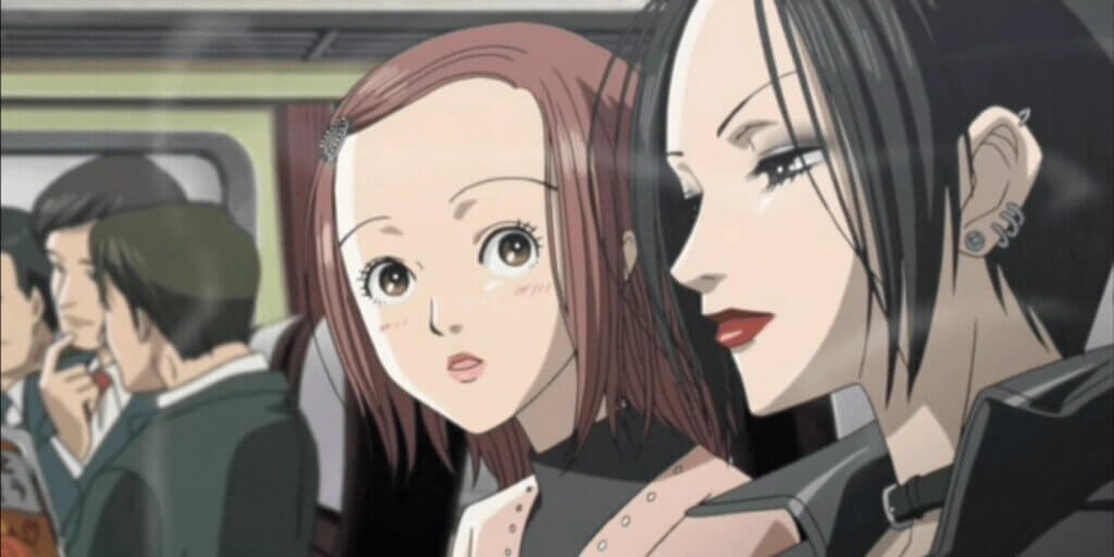 Image is two young women, Nana Komatsu and Nana Osaki. Nana K has short red hair and is wearing a pink sweater over a brown shirt. She is looking at Nana Osaki, who has short black hair, eyeshadow, lipstick, piercings on her left ears, a black choker, and black leather jacket. Some male characters in suits are in the background sitting in train seats.