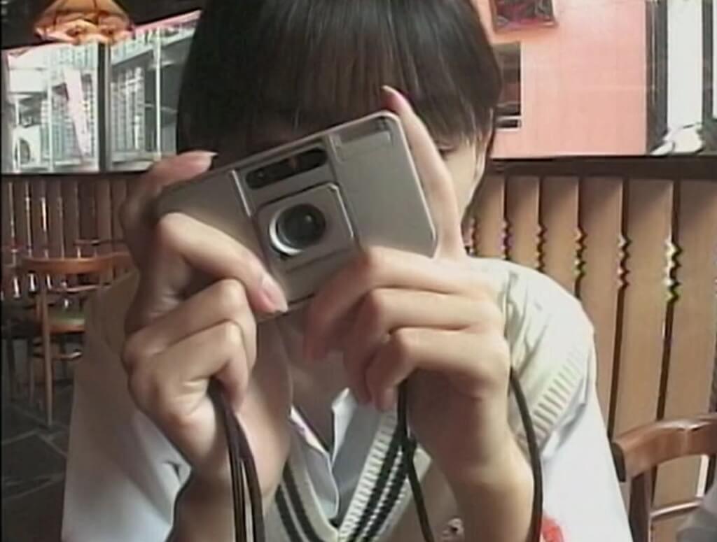 In a cafe, high school girl Hiromi holds a camera in front of her face.