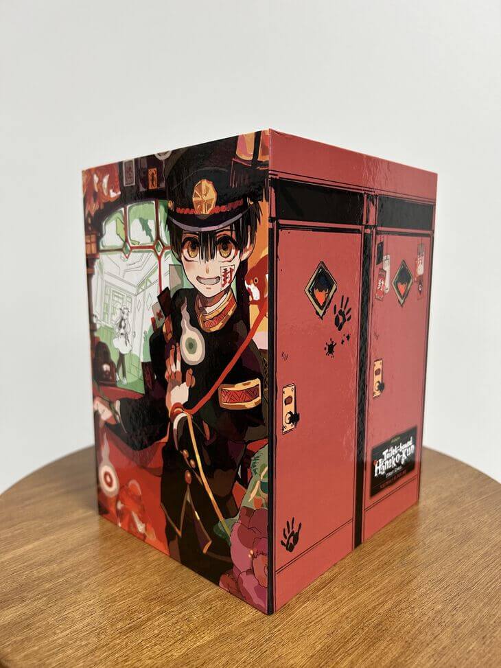The Toilet Bound Hanako-kun box set first installment. It is a small box set designed to look like red high school toilet cubicles marked with hand prints and ink splodges. The side shows a color illustration of a teenage boy wearing an old military style uniform. He is surrounded by will-o-wisps. Through a window in the background is a green and white monotone drawing of a young girl wearing a more modern school uniform.