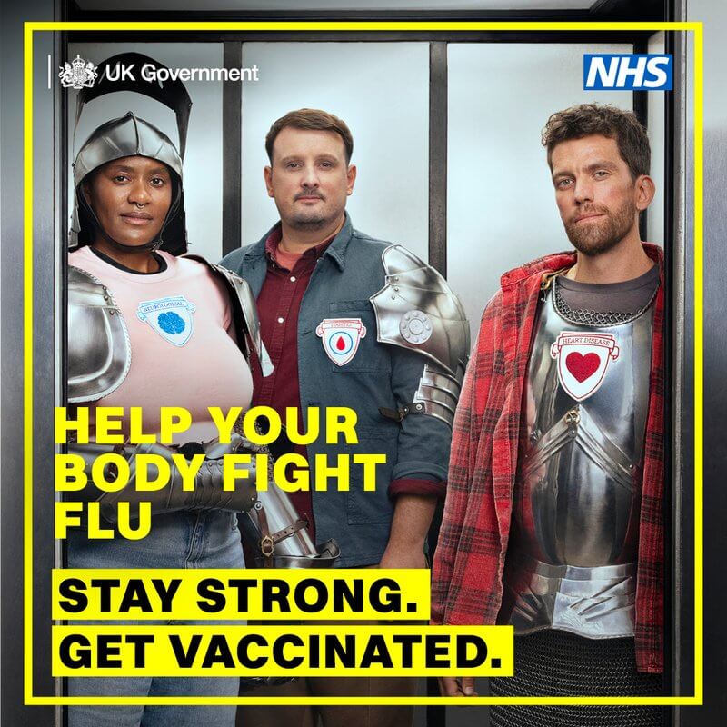 A UK Government NHS public service advertisement. In yellow text it says: “Help your body fight flu”. Underneath that, is black text on a yellow highlighted text says “Stay strong. Get vaccinated.” Three people are in a lift wearing parts of silver armour. A black woman in a pink top has a crest with a blue brain on it with “Neurological” above it. She is wearing a helmet, shoulder braces, and gauntlets. A white man in a blue-grey top has a crest with a blood drop in a circle, with “Diabetes” written above it. Another white man wears a red plaid shirt over the top of a breast plate that has a love heart on it with “Heart Disease” written on it.
