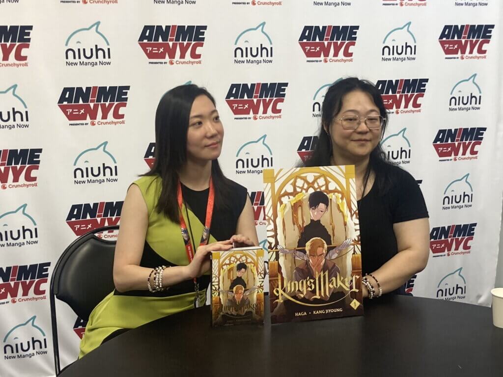 Two women, Haga and Kang Jiyoung, sit while displaying pictures of their comic King's Maker.