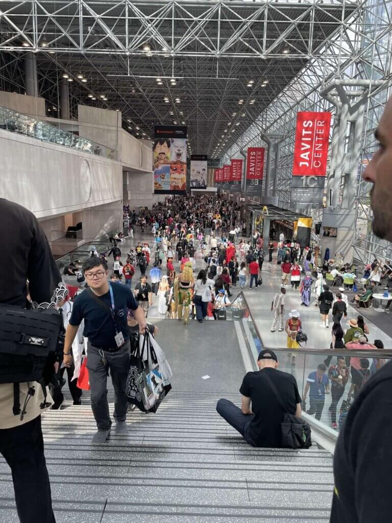Various attendees walk up and down stairs in the Javits Center. One person is sitting on a step.