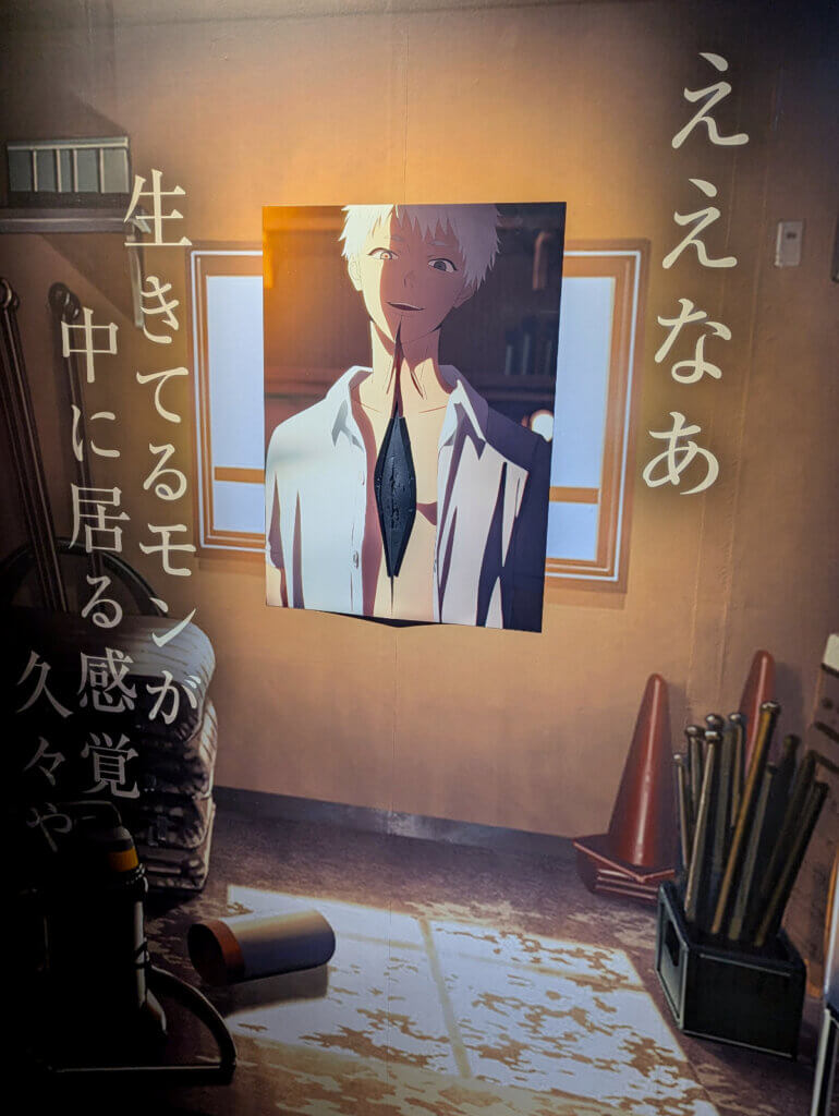 Photograph of a replica scene from The Summer Hikaru Died on a wall featuring a cutout of Hikaru’s face and torso. His torso is split open vertically and there is some kind of black substance covering it.