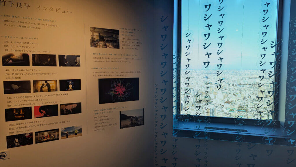 Photograph of a window overlooking buildings with Japanese vertical sound effect text hanging in front of it that appears to be printed on clear plastic. Next to the window is an interview in Japanese with stills from the The Summer Hikaru Died anime.