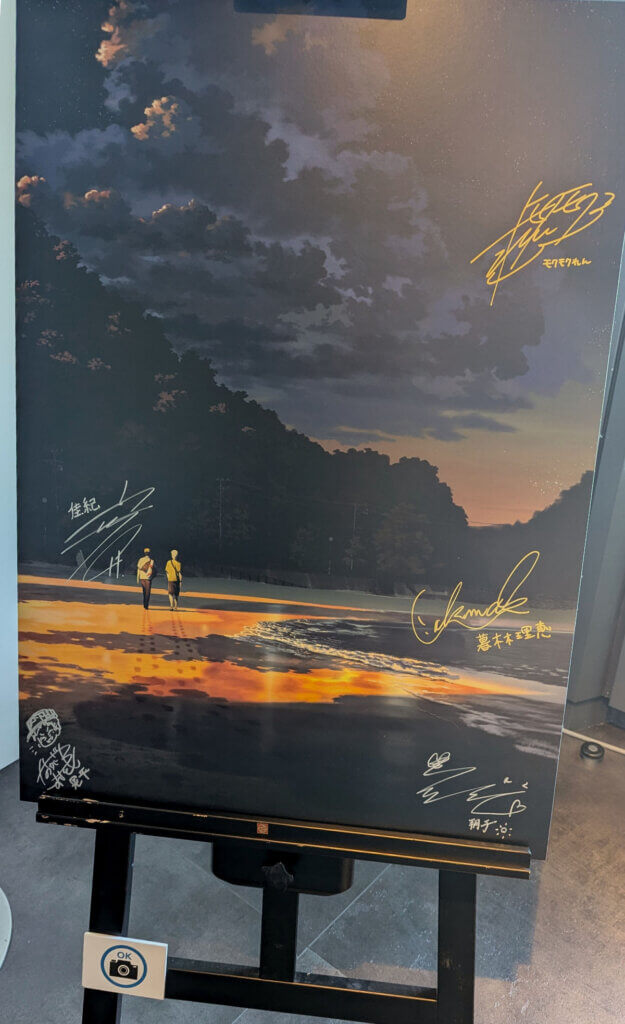 Photo of the The Summer Hikaru Died season 2 visual with staff signatures overlaid in gold and silver pen. Hikaru and Yoshiki are pictured walking away from the viewer on a beach. The sun is setting and there are looming clouds in the sky.