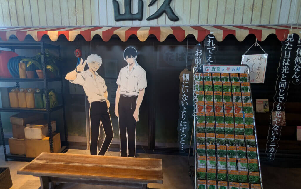 Photograph of a retro Japanese store facade featuring life-size cutouts of Hikaru and Yoshiki from the The Summer Hikaru Died anime. There is a bench in front of them that can be sat on, with a display stand of vegetable seeds next to it.