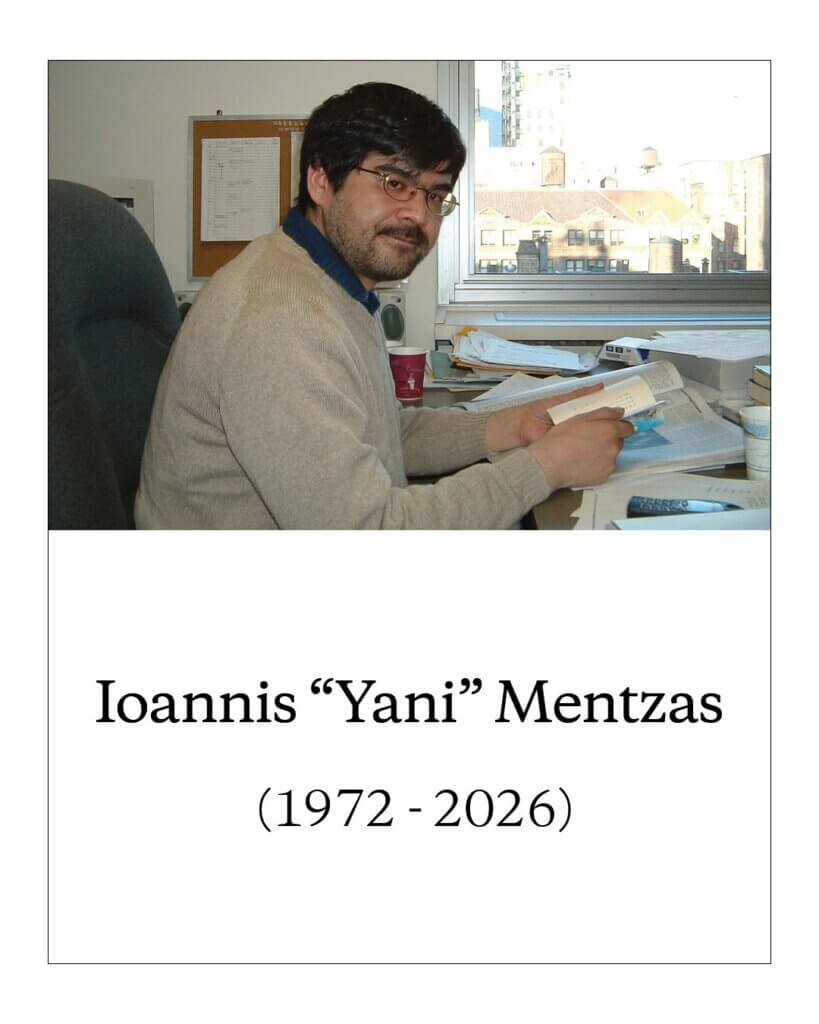 A picture of Ioannis “Yani” Mentzas, marked with the years of his life: 1972-2026. He is wearing a grey sweater and glasses while reading a book at his desk.