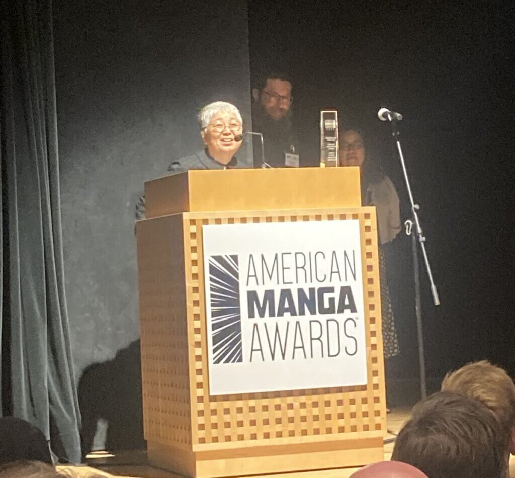 Chigusa Ogino, a woman with short grey hair, speaks at the American Manga Awards podium. Two people can be seen standing behind her.