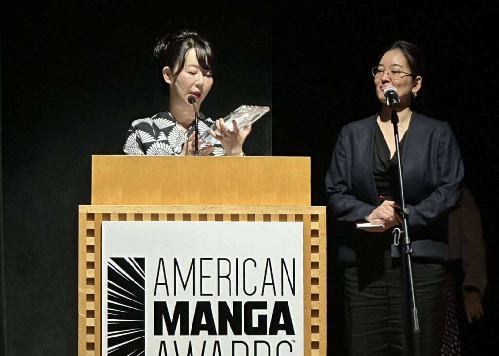 Akiko Higashimura, a woman wearing a white and black kimono, stands at the podium holding her award. An interpreter dressed in black is standing to her right.