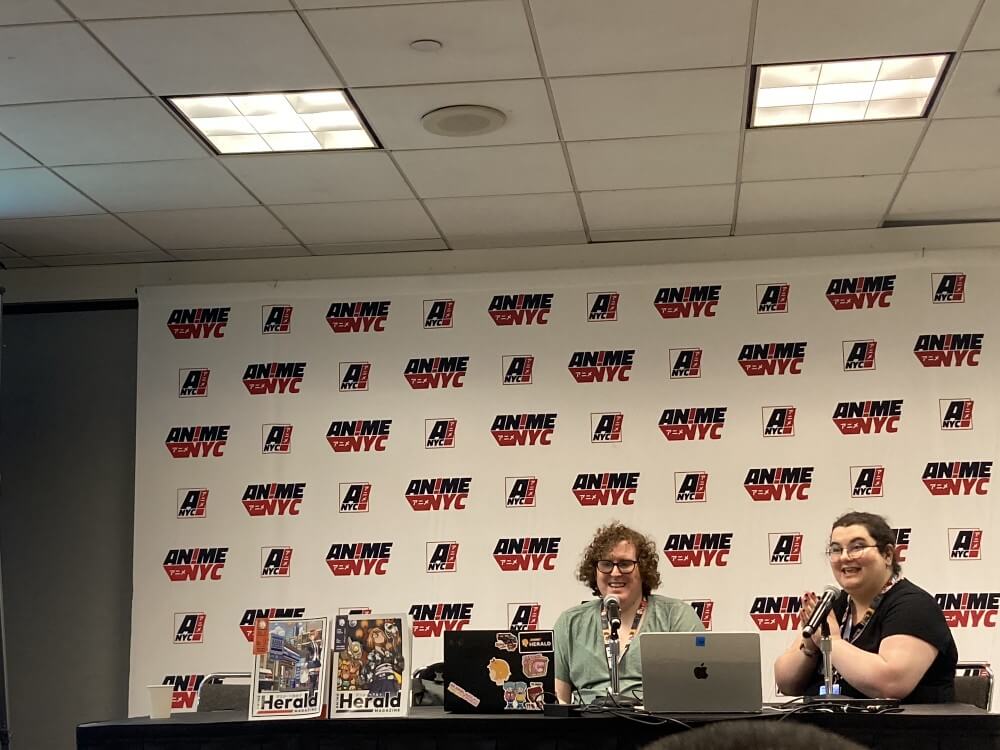 A scene from the Anime Herald Magazine panel. Samantha Ferreira and Katy Castillo sit at a table with copies of Anime Herald Magazine Issues 1 and 2.