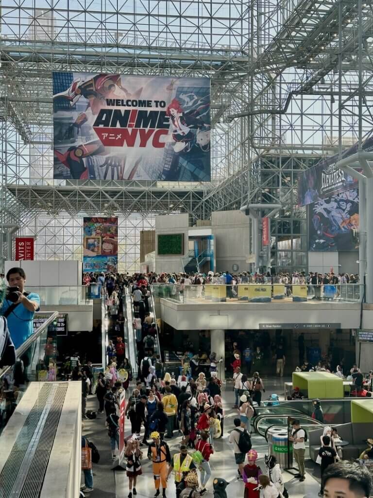 A scene inside the Javits Center during Anime NYC. A large banner reading “Welcome to Anime NYC” is hanging from the ceiling. People are taking the escalator between floors.