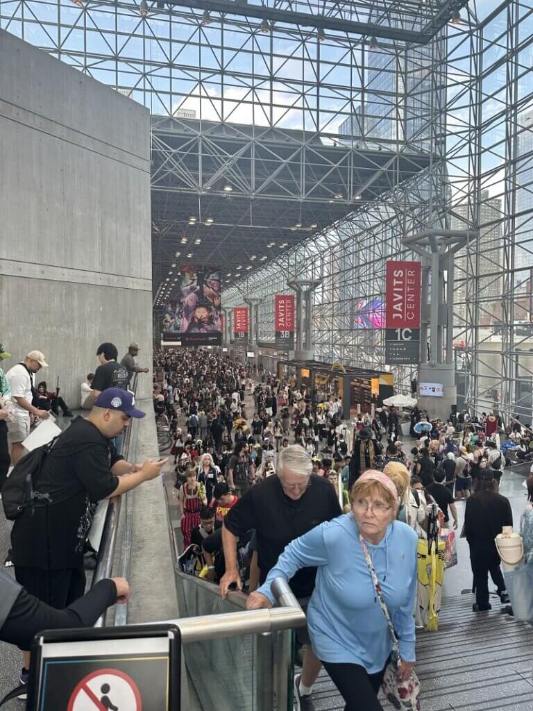 Two older people climb the stairs of the Javits center. An enormous crowd can be seen behind them.