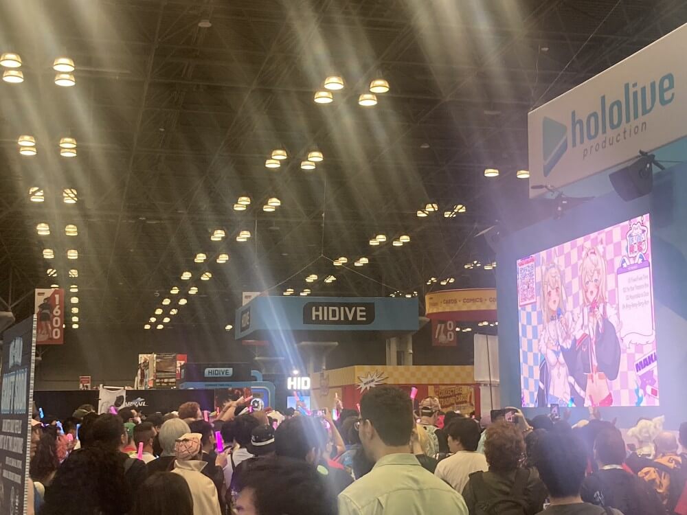 A crowd gathers in front of the hololive booth, where two Vtubers are performing. The banner for Hidive can be seen in the distance.
