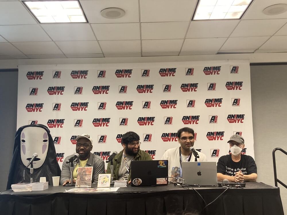A scene from the Manga Mavericks Books panel. Victoria Esnard, Mike Jokoh, Varun Gupta, Siddharth Gupta and Erica Friedman sit together at a table. Esnard is wearing a No Face costume that obscures her body and face.